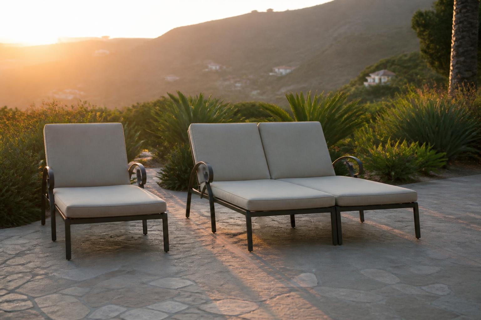 Single chaise lounge beside a double chaise lounge with beige cushions on a stone patio at sunset