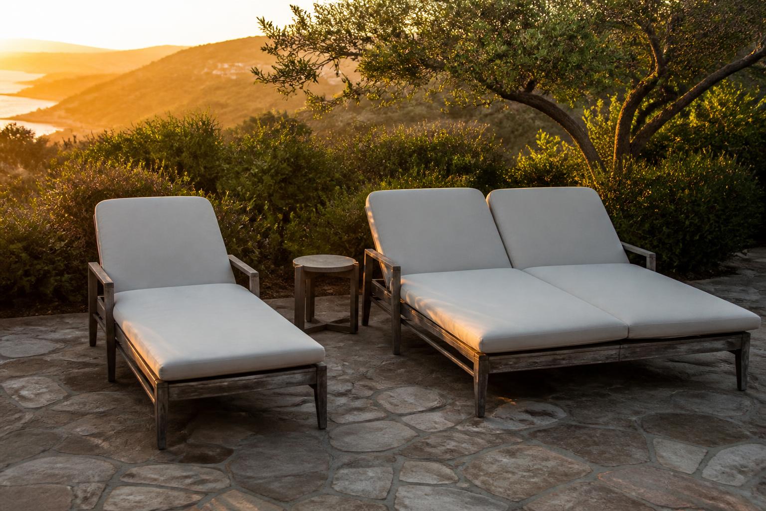 Double chaise lounge and single chaise lounge with light cushions and a small side table on a stone patio at sunset
