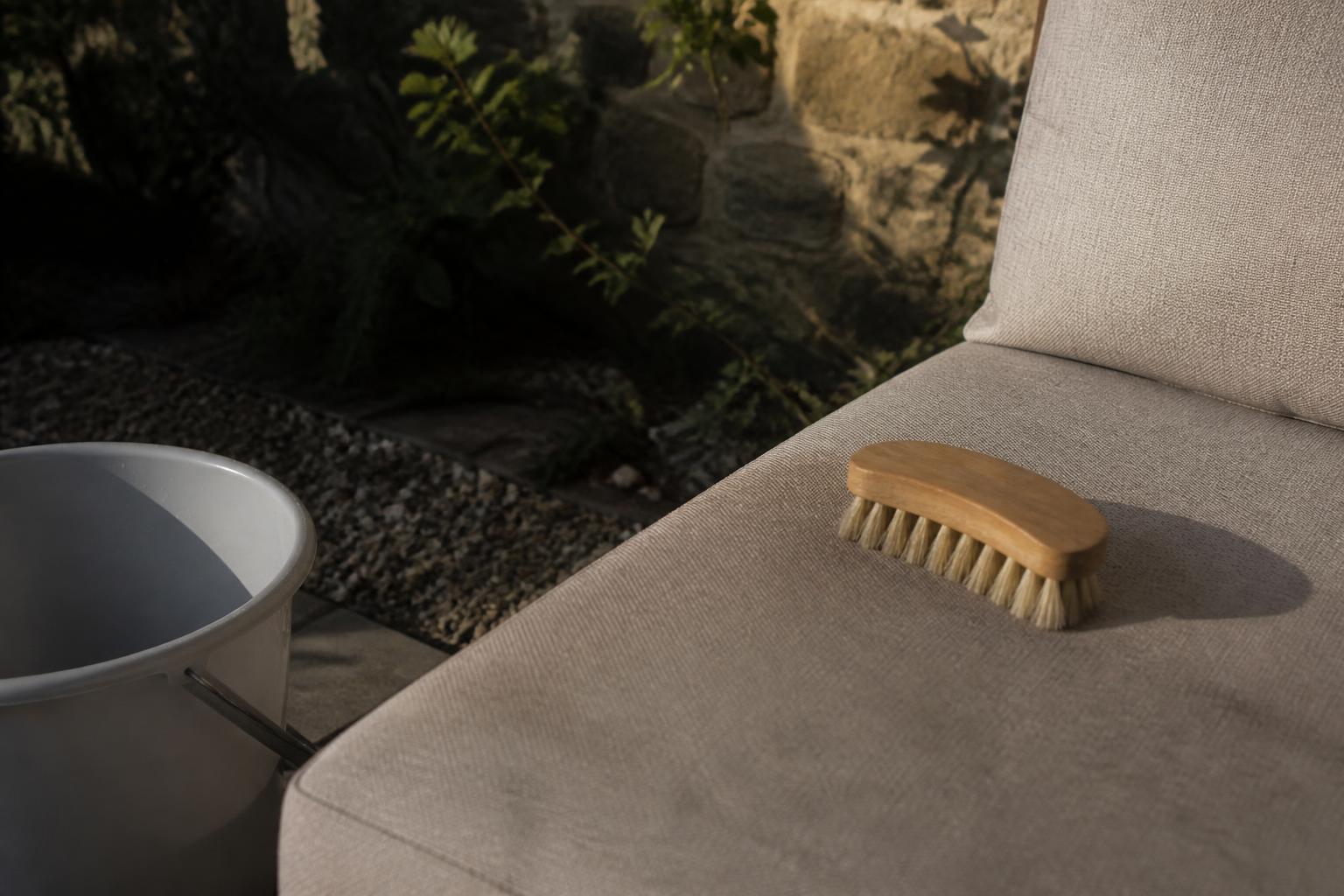 Soft-bristle brush resting on a light gray outdoor cushion beside a white bucket on a patio